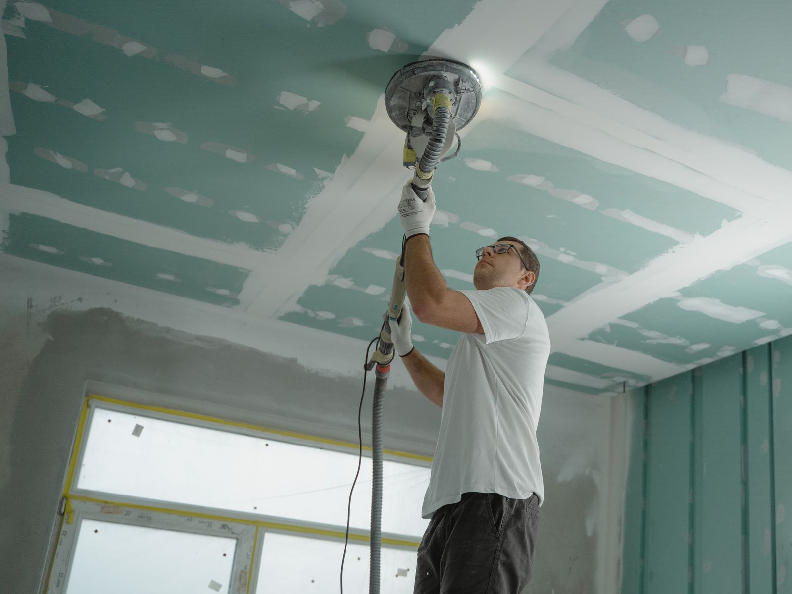 Contractor smoothing a ceiling with a vacuum-attached sander that captures dust at the source