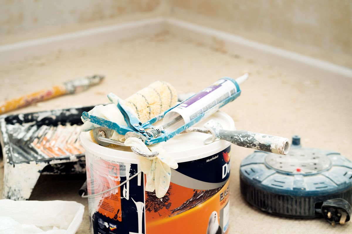 Paint bucket, roller sleeve, and caulking gun staged on protected floors before a ceiling repaint